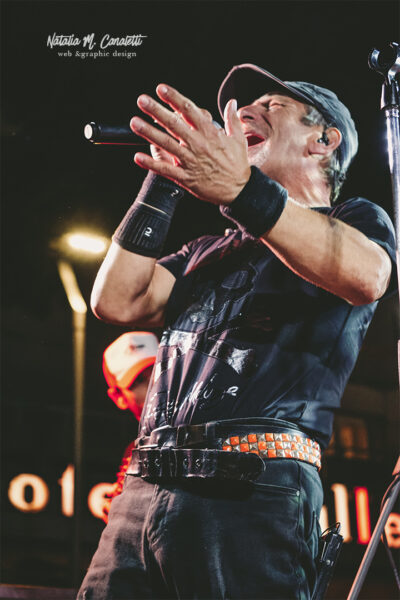 Damiano Busetto Il cantante degli Infrarossi Tribute Band in piena esibizione sul palco, con microfono in mano e mani alzate verso il pubblico, mentre indossa una maglietta nera con un design rock e una cintura con borchie.