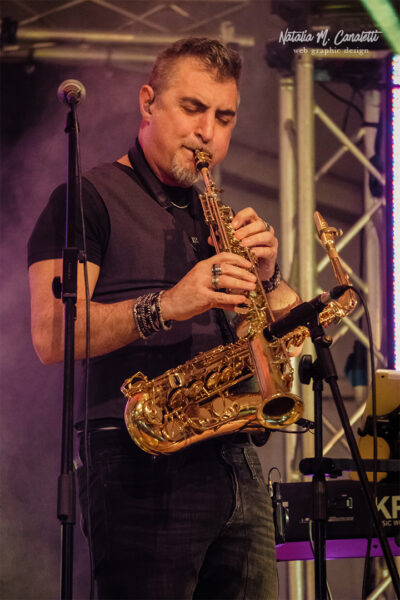Tiziano Valente, sassofonista degli Infrarossi Tribute Band, suona il sax sul palco durante un concerto dal vivo. Indossa un abbigliamento casual con accessori e un'espressione concentrata mentre si esibisce.